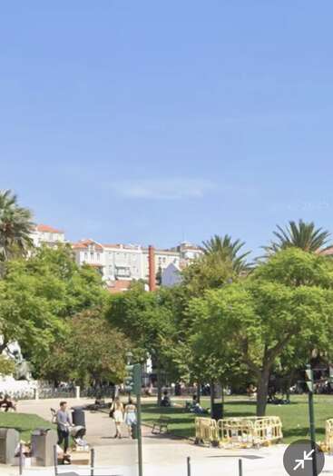 Ein sonniger Stadtpark mit grünen Bäumen, Menschen, die spazieren gehen und sich auf Bänken entspannen, umgeben von Gebäuden mit roten Dächern unter einem klaren blauen Himmel. Friedliche Atmosphäre.