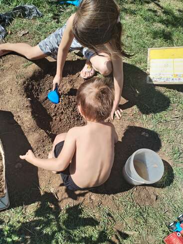 Zwei Kinder spielen draußen in der Sonne und graben mit einer blauen Schaufel in der Erde. Ein Plastikeimer und ein Buch liegen in der Nähe auf dem Gras.