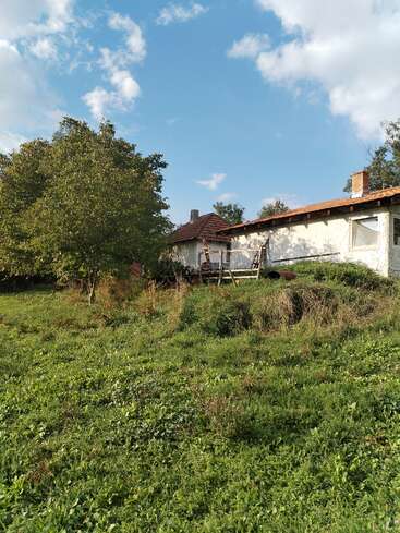 Ein rustikales Haus mit rotem Ziegeldach steht umgeben von saftigem, grünem Gras und einem Baum. Der Himmel darüber ist hell und teilweise bewölkt.