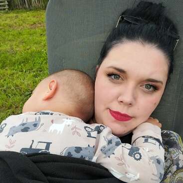 Une femme aux cheveux foncés et au rouge à lèvres rouge berce un bébé endormi dans ses bras. Elles semblent détendues, assises dehors sur de l'herbe verte.