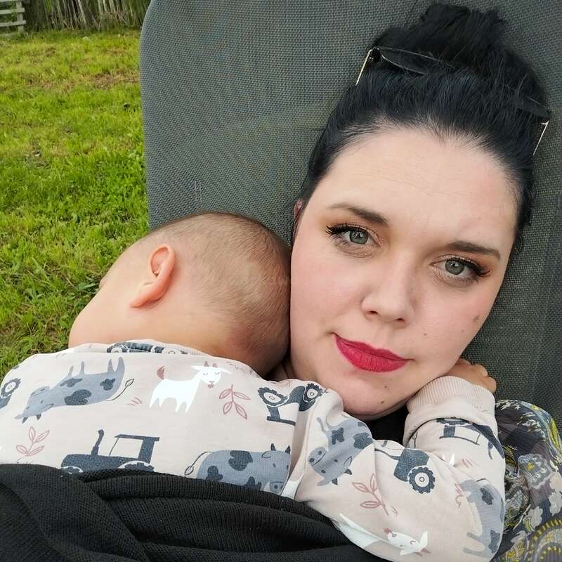 Eine Frau mit dunklem Haar und rotem Lippenstift wiegt ein schlafendes Baby in ihren Armen. Sie wirken entspannt und sitzen draußen im grünen Gras.