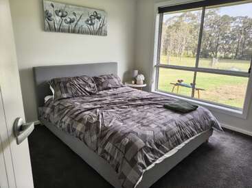 Um quarto moderno e aconchegante com uma cama estofada cinza, roupa de cama geométrica, mesa de cabeceira, uma grande janela com vista para campos verdes e uma obra de arte floral acima da cabeceira.
