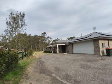Uma casa moderna de tijolos com uma grande garagem fica em uma propriedade rural com uma entrada de automóveis de cascalho, cercada por árvores, vegetação e uma cerca de madeira preta.
