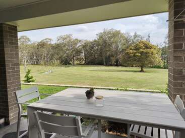 Um pátio coberto com uma mesa e cadeiras brancas tem vista para um gramado verde espaçoso, cercado por árvores e céu aberto, criando um ambiente externo tranquilo e relaxante.