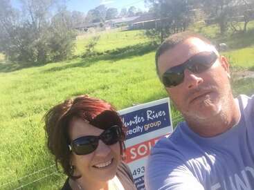 Um casal feliz posa para uma selfie em frente a uma placa "Hunter River Realty Group" onde se lê "VENDIDO" em uma propriedade gramada e ensolarada.