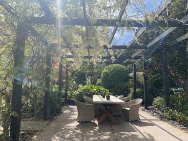 Sonnenlicht fällt auf eine Gartenterrasse mit einem Holztisch und Korbstühlen unter einer begrünten Pergola, umgeben von üppigem Grün und blühenden Glyzinien.