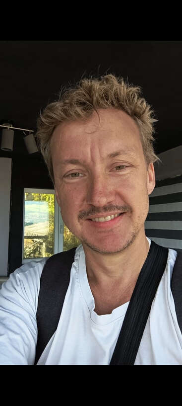 Un homme aux cheveux blonds bouclés, à la barbe légère et vêtu d'une chemise blanche sourit pour un selfie en intérieur. Une lumière naturelle douce pénètre par la fenêtre derrière lui.