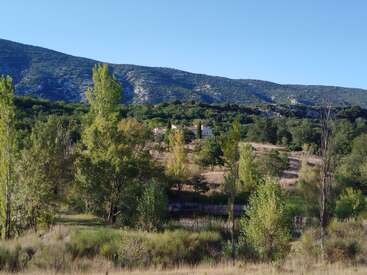 Una encantadora casa enclavada entre frondosos árboles verdes y suaves colinas, con un telón de fondo de escarpadas montañas bajo un cielo azul claro y brillante.