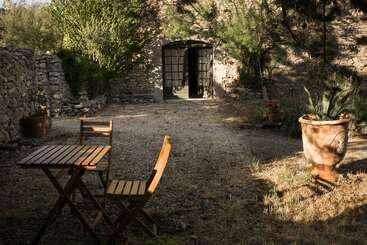 Un patio rústico al aire libre con un muro de piedra, mesa y sillas de madera, una gran maceta, vegetación y la luz del sol proyectando sombras cerca de una puerta arqueada.