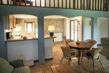 Este acogedor espacio rústico cuenta con paredes de piedra, una pequeña cocina, una mesa de comedor redonda de madera con sillas de mimbre, iluminación cálida, suelo de baldosas y una barandilla de madera en la escalera.