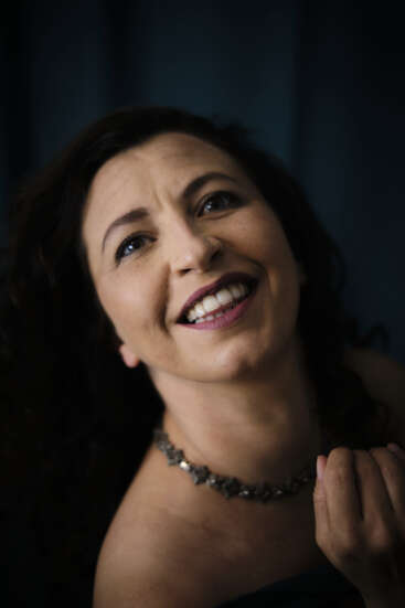 Una mujer de pelo oscuro ondulado sonríe cariñosamente a la cámara. Lleva un collar oscuro, un maquillaje sutil y posa sobre un fondo suave y oscuro.