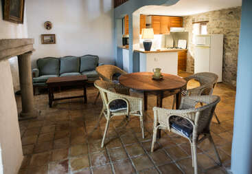 Este acogedor salón cuenta con una mesa redonda de madera con sillas de mimbre, un sofá verde, una chimenea rústica, suelo de baldosas y una pequeña cocina con armarios de madera.