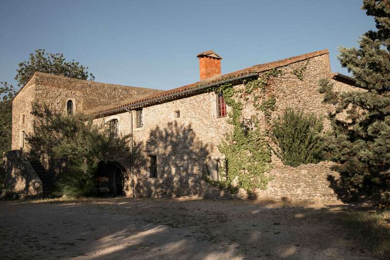 The image depicts a rustic stone building with a red-tiled roof, adorned with green vines, set against a clear blue sky on a sunny day.