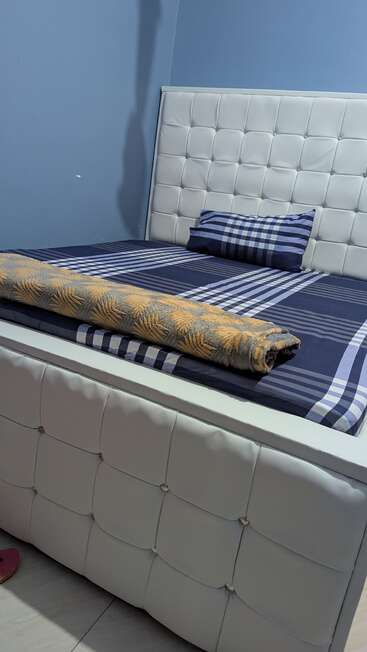 Esta imagem mostra uma cama moderna bem arrumada com cabeceira e base brancas tufadas, roupa de cama xadrez azul, um travesseiro combinando e um cobertor com padrão enrolado.
