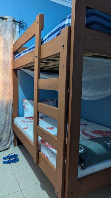 Esta imagem mostra um beliche de madeira em um quarto com paredes azuis. A cama inferior tem roupas de cama com estampas de corações e um cobertor. Os chinelos azuis estão no piso de ladrilhos.