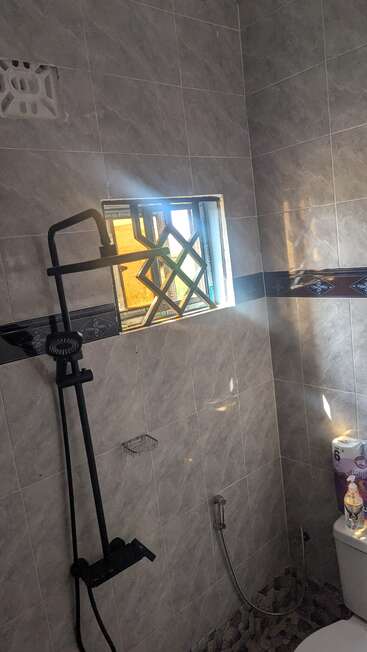Um banheiro com paredes de azulejos cinzas, um chuveiro preto, uma pequena janela aberta com barras de metal, vaso sanitário, pacote de papel higiênico e luz do sol entrando, criando sombras.