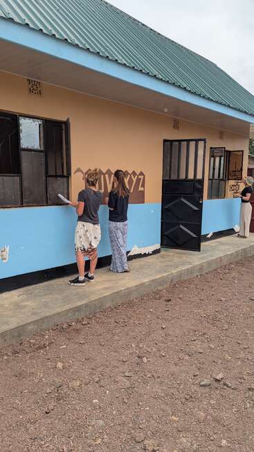 Três pessoas pintam murais educativos na parte externa de um prédio bronzeado e azul, provavelmente uma escola. O chão é rochoso; a atmosfera parece calma e produtiva.