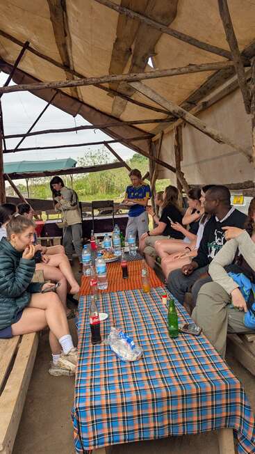 Um grupo de pessoas senta-se ao redor de uma mesa sob um abrigo rústico, compartilhando bebidas, lanches e conversas, desfrutando de uma reunião casual em um ambiente natural ao ar livre.