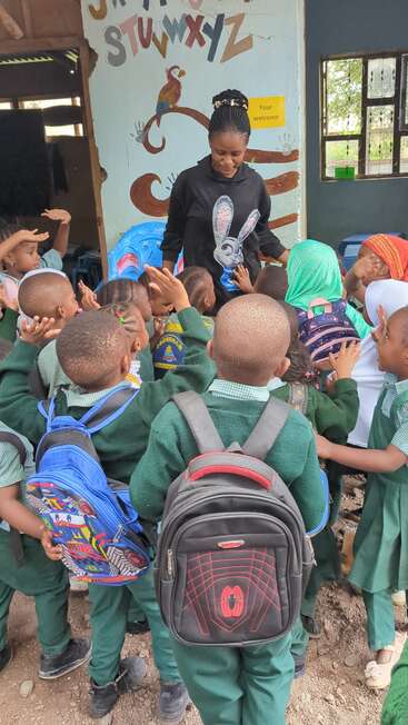 A group of young children in green uniforms eagerly surround a smiling teacher. Colorful backpacks, alphabet wall art, and open hands create a lively classroom scene.