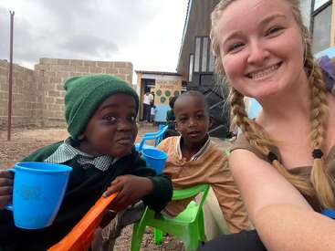 A woman with braided hair smiles while taking a selfie with two young children sitting on colorful chairs, one drinking from a blue cup, outdoors.
