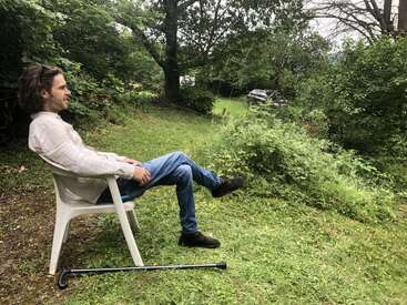 Ein Mann sitzt entspannt auf einem weißen Plastikstuhl im Freien, umgeben von Grünzeug. Ein schwarzer Stock ruht vor ihm auf dem Boden, friedliche Atmosphäre.