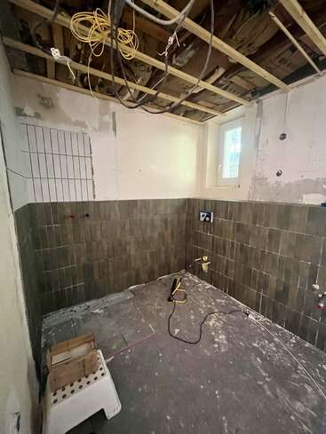 This image shows a room under renovation with exposed ceiling beams, hanging wires, unfinished walls, partially tiled walls, and construction materials scattered on the dusty floor.
