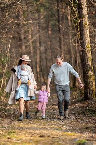 Una familia de cuatro miembros, compuesta por los padres y dos niños pequeños, camina alegremente por un sendero en el bosque, rodeada de árboles altos y disfrutando juntos de la naturaleza.