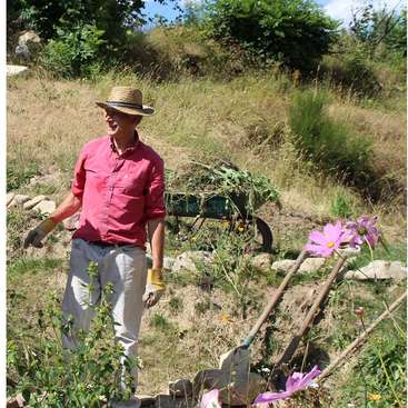 Ein Mann mit rotem Hemd und Strohhut steht in einem Garten, umgeben von Blumen und einer Schubkarre, trägt Handschuhe und hält ein Gartengerät.