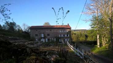 Une maison rustique en pierre avec un toit en tuiles rouges se trouve au bout d'une route de campagne, entourée d'arbres, de verdure et d'un paysage rural paisible.