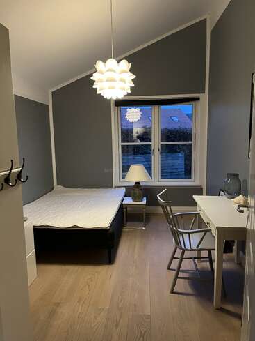 Un dormitorio moderno y minimalista con paredes grises, suelo de madera, una cama individual, un escritorio blanco, una silla de madera, una lámpara de mesa, una elegante lámpara de techo y una gran ventana.