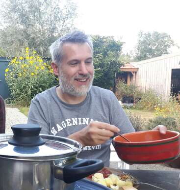 Un hombre sonriente con el pelo canoso disfruta de una comida al aire libre. Sostiene un cuenco rojo y una cuchara, rodeado de comida, flores, vegetación y un cobertizo de madera al fondo.