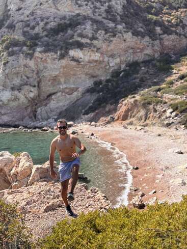 Ein Mann ohne Hemd, in Shorts und mit Sonnenbrille klettert auf felsigem Gelände in der Nähe eines abgelegenen Strandes mit klarem türkisfarbenem Wasser, umgeben von schroffen Klippen und üppigem Grün.