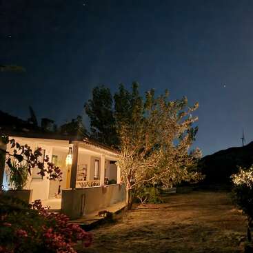 Ein gemütliches Haus mit einer Veranda ist nachts sanft beleuchtet. Bäume und Pflanzen umgeben das Haus, während die Sterne am klaren, dunklen Himmel über ihm glitzern.
