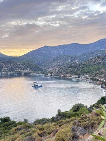 Eine ruhige Bucht, umgeben von üppigen Hügeln, mit Yachten, die in ruhigem Wasser unter einem wolkenverhangenen Himmel bei Sonnenuntergang ankern und eine friedliche und malerische Küstenszene schaffen.