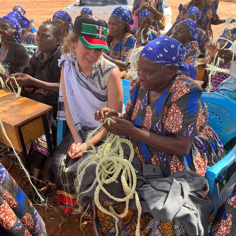 The image depicts a group of people, predominantly women, gathered in a circle, engaged in a craft activity, with a white car and trees visible in the background.