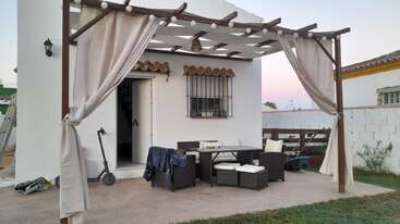 Eine gemütliche Terrasse hat eine Holzpergola mit Vorhängen, Lichterketten, Korbmöbeln, einem Tisch und einem Roller, die neben einem weißen Haus stehen.