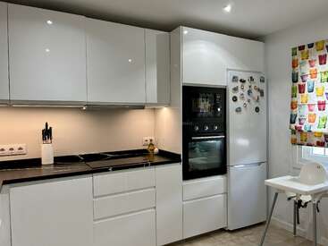 This modern kitchen features white cabinets, a black countertop, built-in oven, refrigerator with magnets, a knife set, colorful window curtain, and a white high chair.