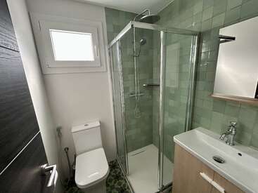 This compact bathroom features a glass shower enclosure, green tiled walls, a modern toilet, sink with cabinet, mirror, small window, and patterned floor tiles.