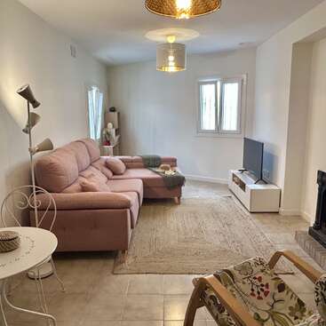 This cozy living room features a pink sectional sofa, light rug, TV, lamp, wooden chair with floral cushion, small table, and minimalistic decor in neutral tones.