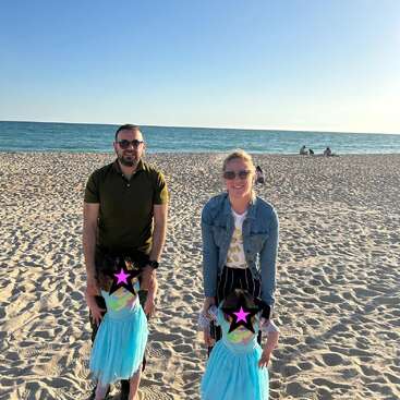 A man and woman with two young girls in blue dresses stand on a sandy beach. The ocean and blue sky are in the background.