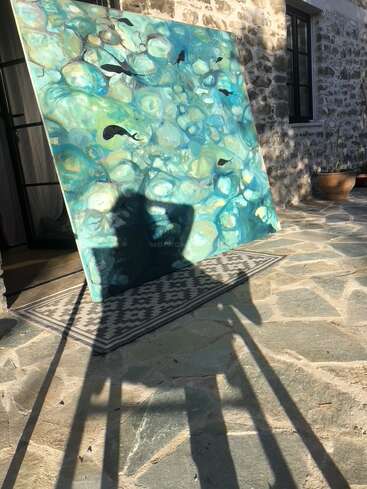 Ein großes, farbenfrohes Gemälde, das eine Unterwasserszene mit Fischen zeigt, lehnt an einer Steinmauer und wirft lange Schatten auf eine sonnenbeschienene Steinterrasse.