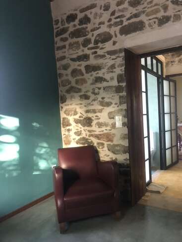 Ein kastanienbrauner Sessel steht neben einer rustikalen Steinmauer und einer blaugrünen Wand. Sonnenlicht fällt herein und wirft Schatten. Eine offene Tür gibt den Blick auf einen weiteren Raum dahinter frei.