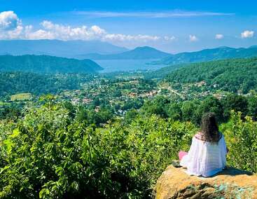 Une femme est assise sur un rocher, contemplant une magnifique vallée verdoyante parsemée de maisons, de forêts denses, de montagnes et d’un lac bleu paisible, sous un ciel éclatant.
