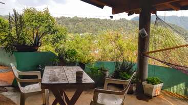 Une terrasse chaleureuse est agrémentée d'une table rustique en bois, de deux chaises en plastique, d'un hamac, de plantes en pot luxuriantes et offre une vue panoramique sur les montagnes verdoyantes en arrière-plan.