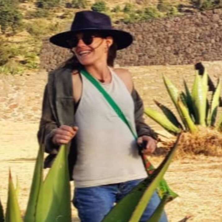 Une personne souriante, coiffée d'un chapeau, portant des lunettes de soleil, un débardeur et un jean, se tient à l'extérieur au milieu d'agaves, avec des collines et des arbres en arrière-plan sous un soleil radieux.