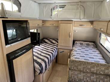 This image shows the interior of a camper van with two single beds, overhead cabinets, a wardrobe, a fridge, a microwave, and minimalistic light wood decor.