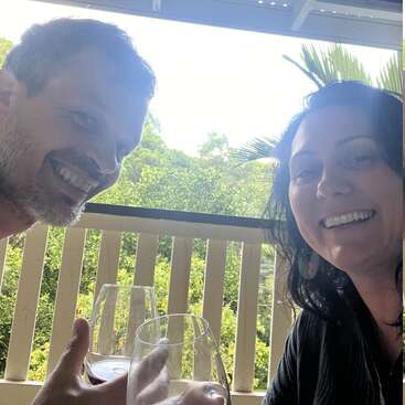 A smiling man and woman sit on a balcony, holding glasses of wine, enjoying a bright, lush, green outdoor view, sharing a cheerful, happy moment together.