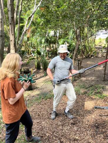 Two people are outdoors in a garden or forest area, smiling and laughing. One person holds a rake, the other holds a green plush toy, both seem happy.