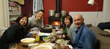 Four people are sitting around a table enjoying a meal together, smiling warmly. Behind them, a cozy fireplace is burning, creating a friendly, inviting atmosphere.