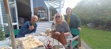 Three people enjoy a casual outdoor gathering in a garden, smiling near a wooden table with food, drinks, and a dog. The atmosphere appears relaxed and friendly.